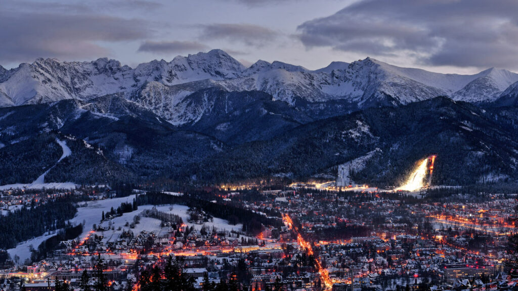 Noclegi Zakopane – widok na rozświetlone nocą miasto na tle ośnieszonych szczytów Tatr